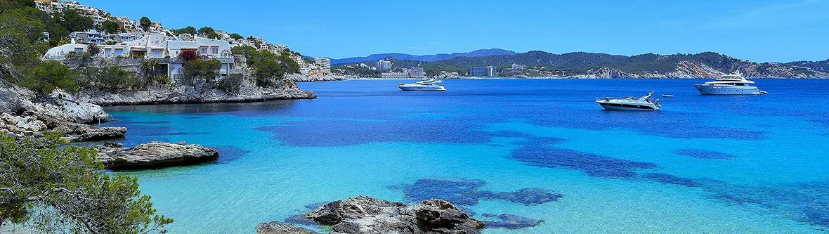 Panoramablick auf eine türkisblaue Mittelmeerbucht in Mallorca mit weißen Yachten, felsiger Küstenlinie und weißen Häusern am Hang